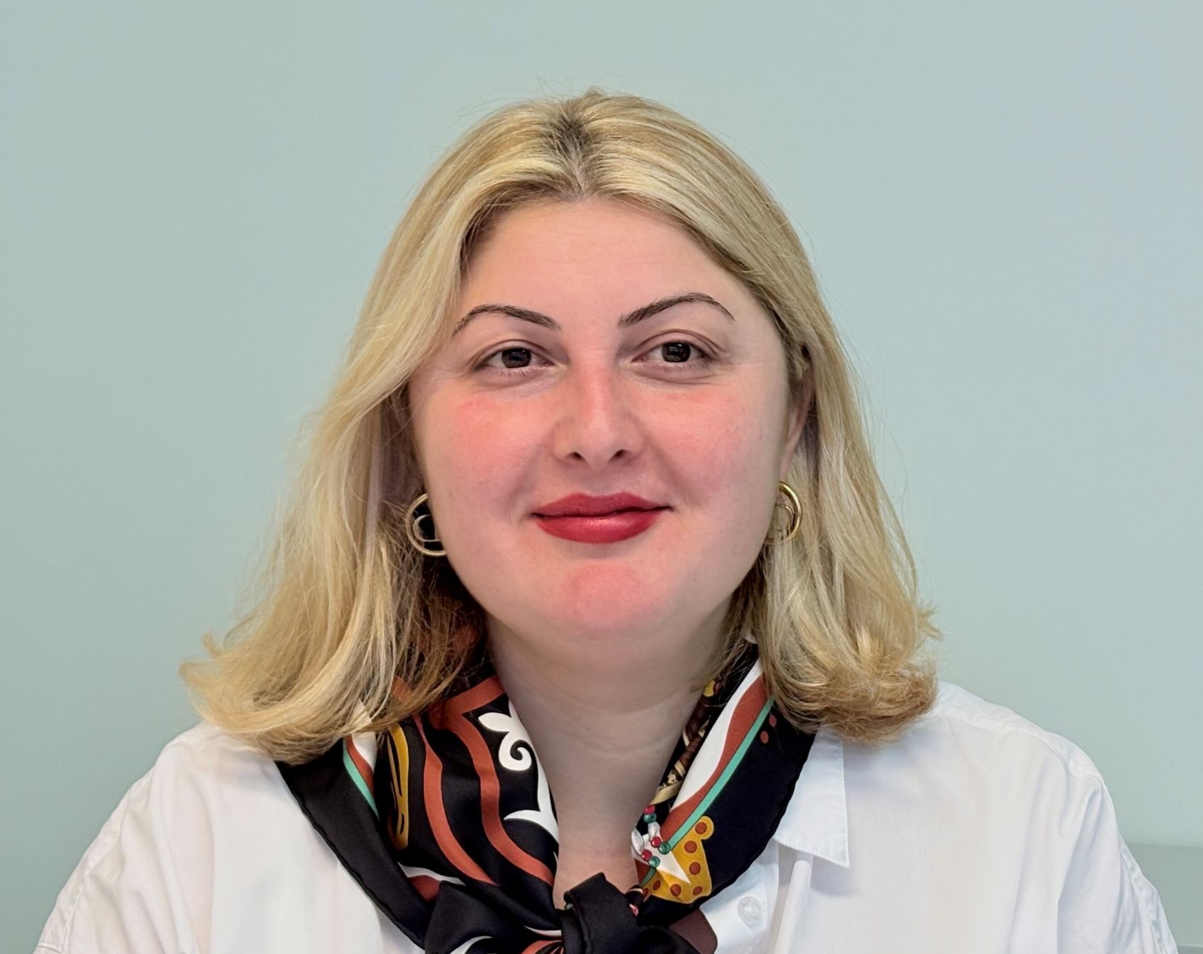 Frau mit blondem Haar, freundlichem Gesichtsausdruck, rotem Lippenstift und gemustertem Halstuch